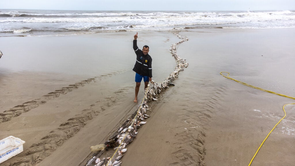 Na praia, pescador mostra rede cheia de peixes. Ele veste casaco e está com um dos braços levantados